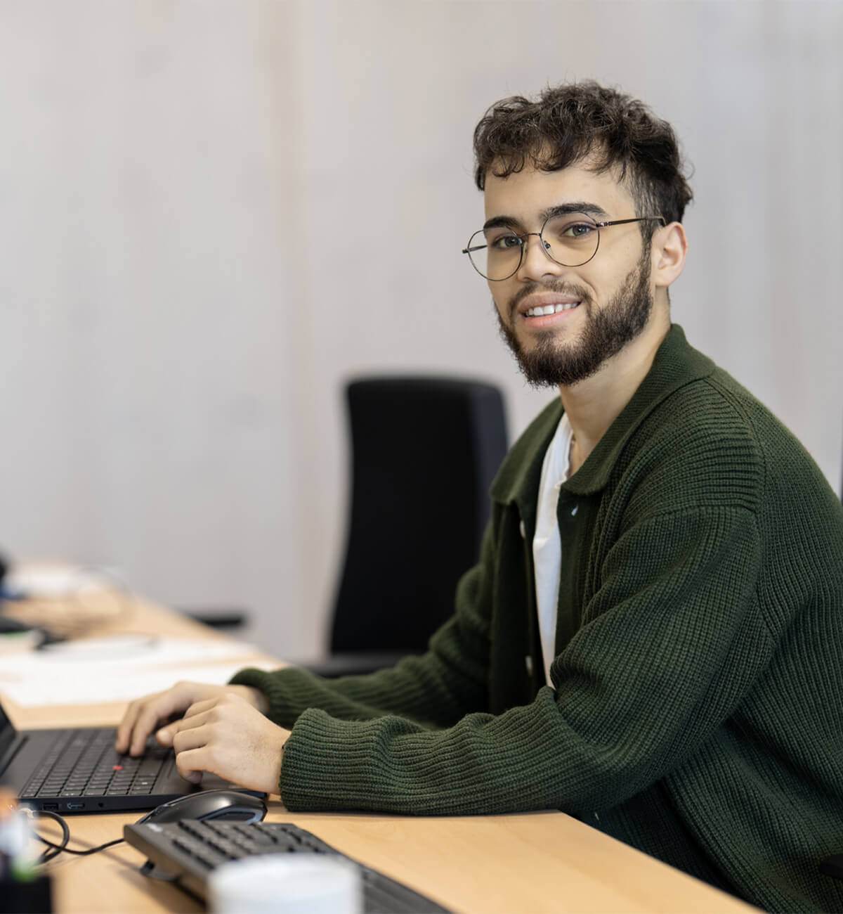 Junger Mann mit Brille und Bart sitzt lächelnd an einem Schreibtisch und tippt auf einer Tastatur und schaut zur Kamera