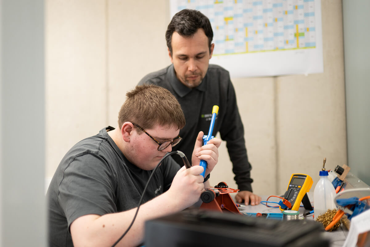 Eine Person arbeitet konzentriert an einem elektronischen Bauteil an einem Arbeitstisch mit Messgeräten und Werkzeugen, der Ausbilder steht daneben