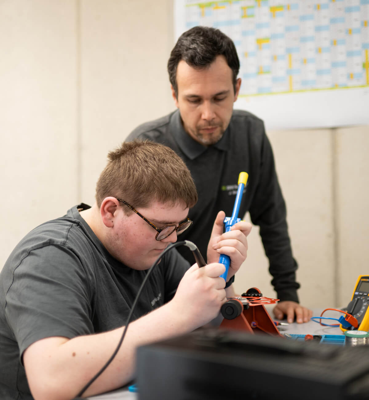 Eine Person arbeitet konzentriert an einem elektronischen Bauteil an einem Arbeitstisch mit Messgeräten und Werkzeugen, der Ausbilder steht daneben