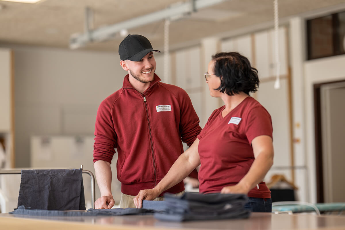 Zwei Personen in roten Oberteilen stehen an einem Tisch mit gefalteter Wäsche und unterhalten sich
