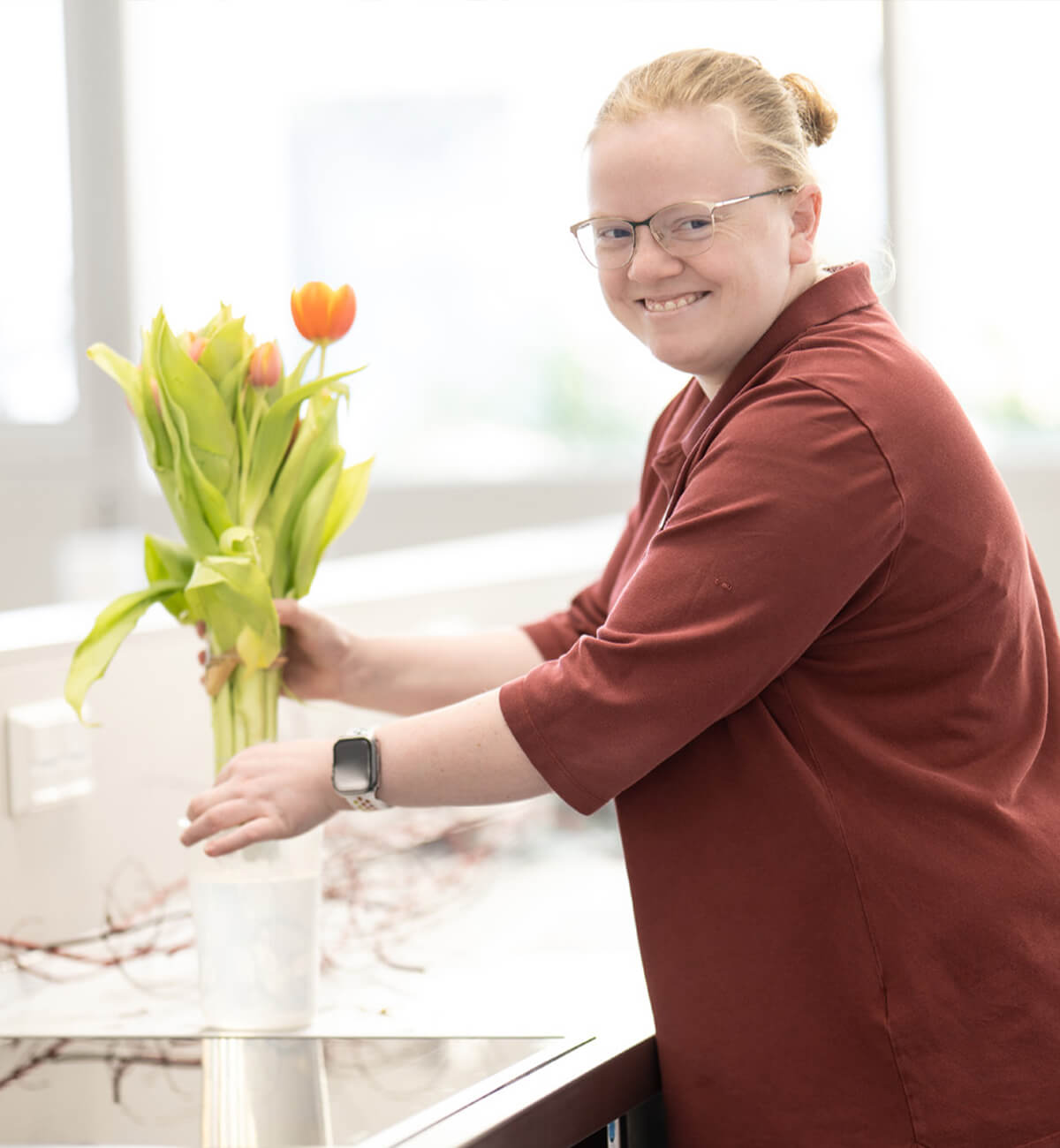 Lächelnde Frau mit Brille und rotem Oberteil stellt Tulpen in eine Vase vor einer Küchenarbeitsplatte