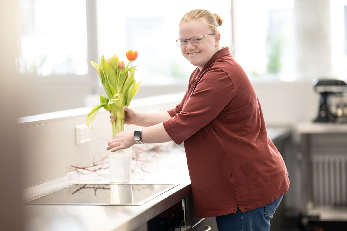 Lächelnde Frau mit Brille und rotem Oberteil stellt Tulpen in eine Vase vor einer Küchenarbeitsplatte