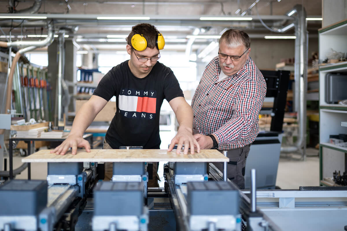Ein junger Mann mit Gehörschutz und ein Ausbilder mit Brille prüfen gemeinsam ein Holzbrett in einer Werkstatt mit Maschinen im Hintergrund