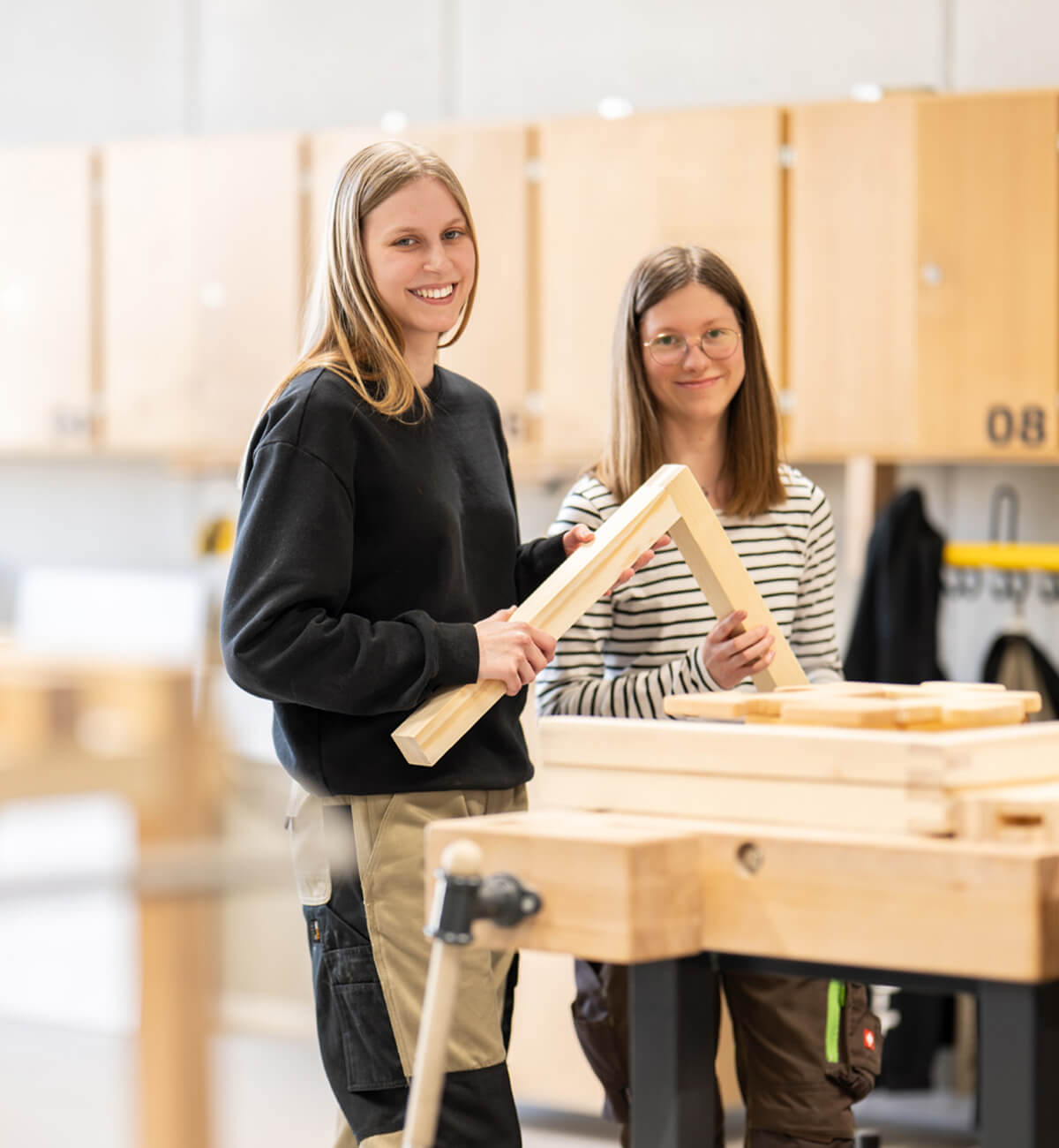Zwei junge Frauen in einer Werkstatt halten ein Holzrahmenstück und lächeln in die Kamera
