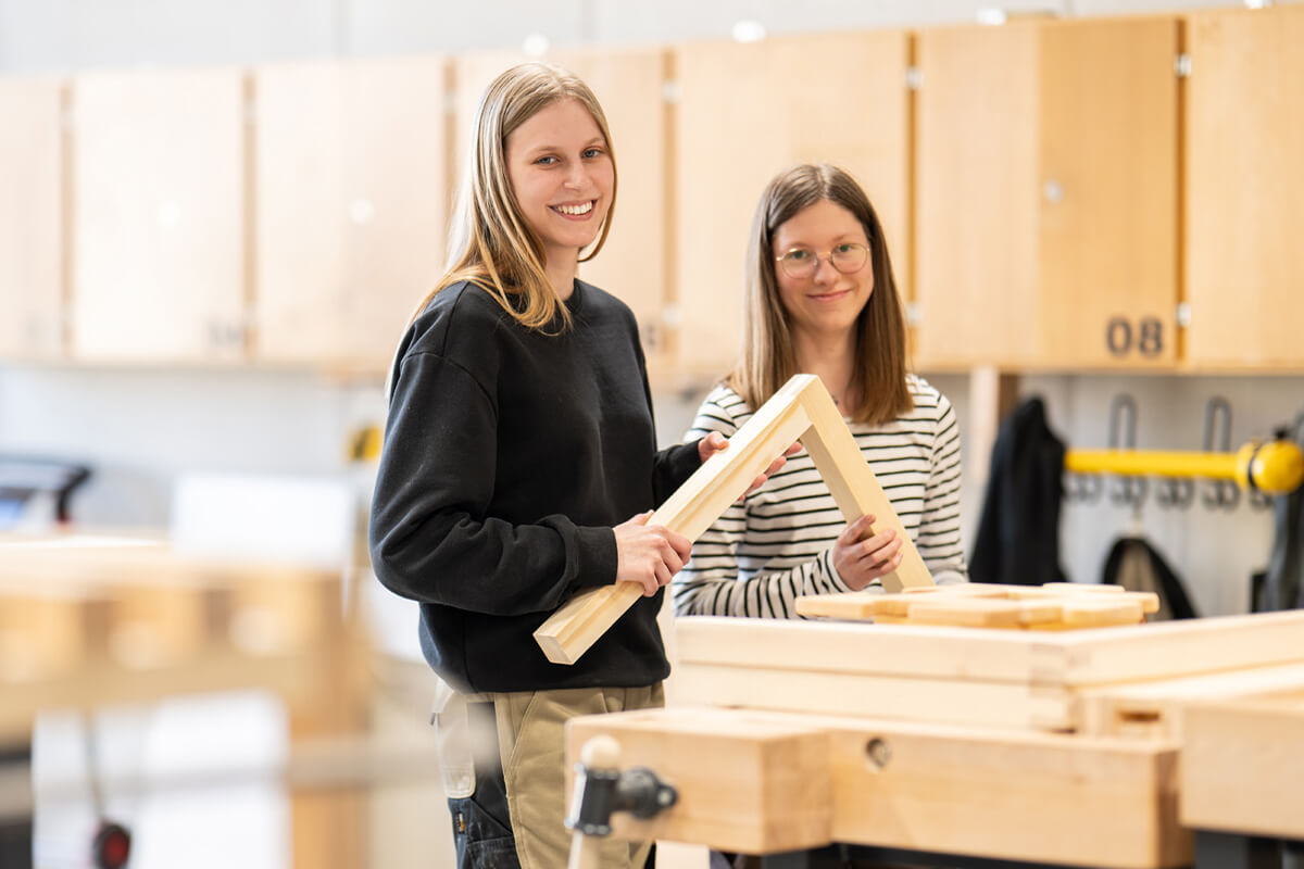 Zwei junge Frauen in einer Werkstatt halten ein Holzrahmenstück und lächeln in die Kamera