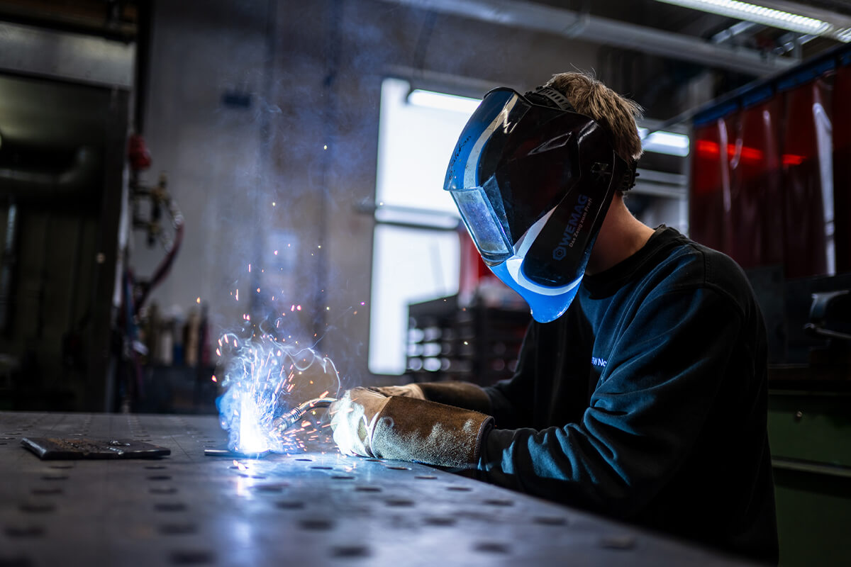 Person mit Schutzhelm und Handschuhen beim Schweißen an einer Metallplatte in einer Werkstatt