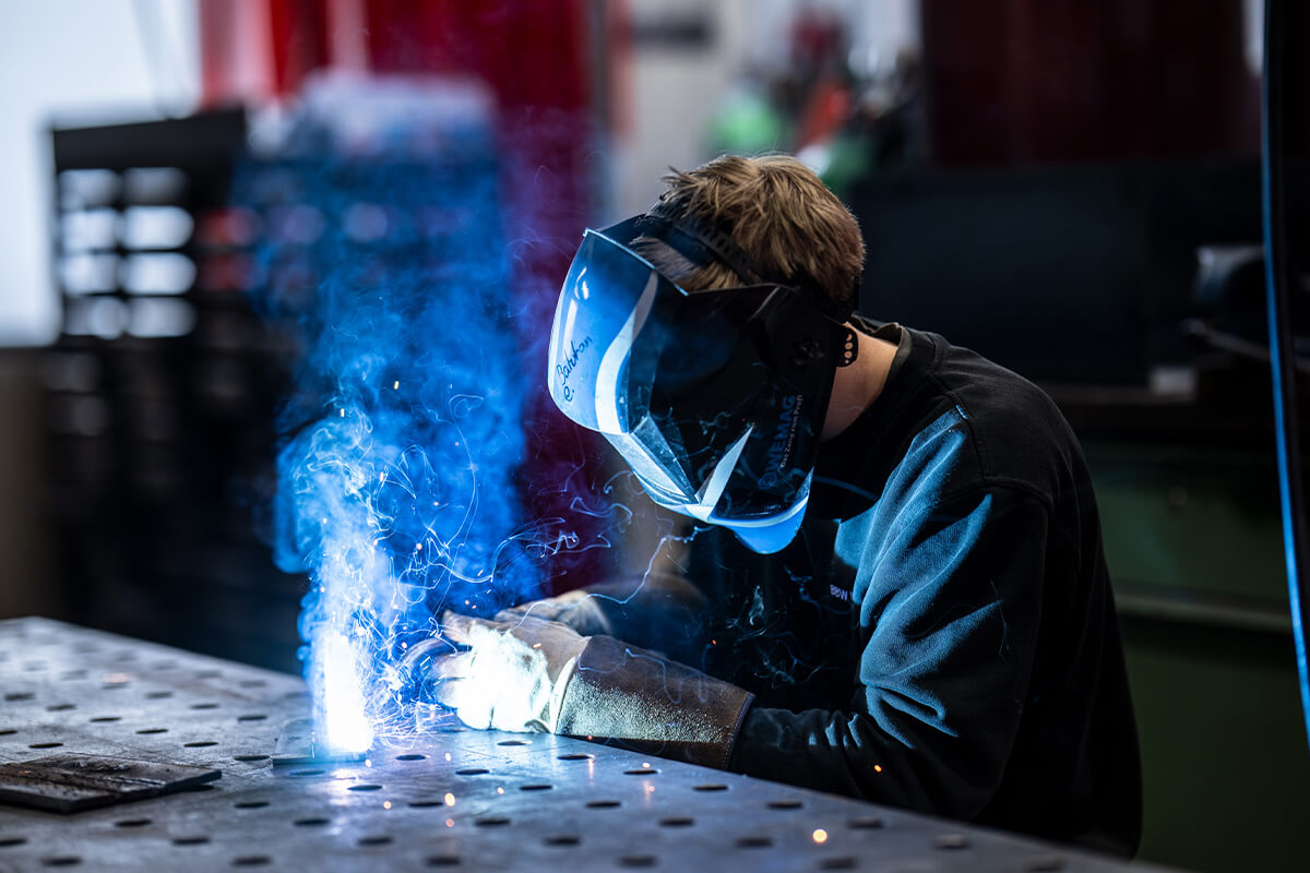 Person mit Schutzhelm und Handschuhen beim Schweißen an einem Metalltisch mit Funken und Rauch