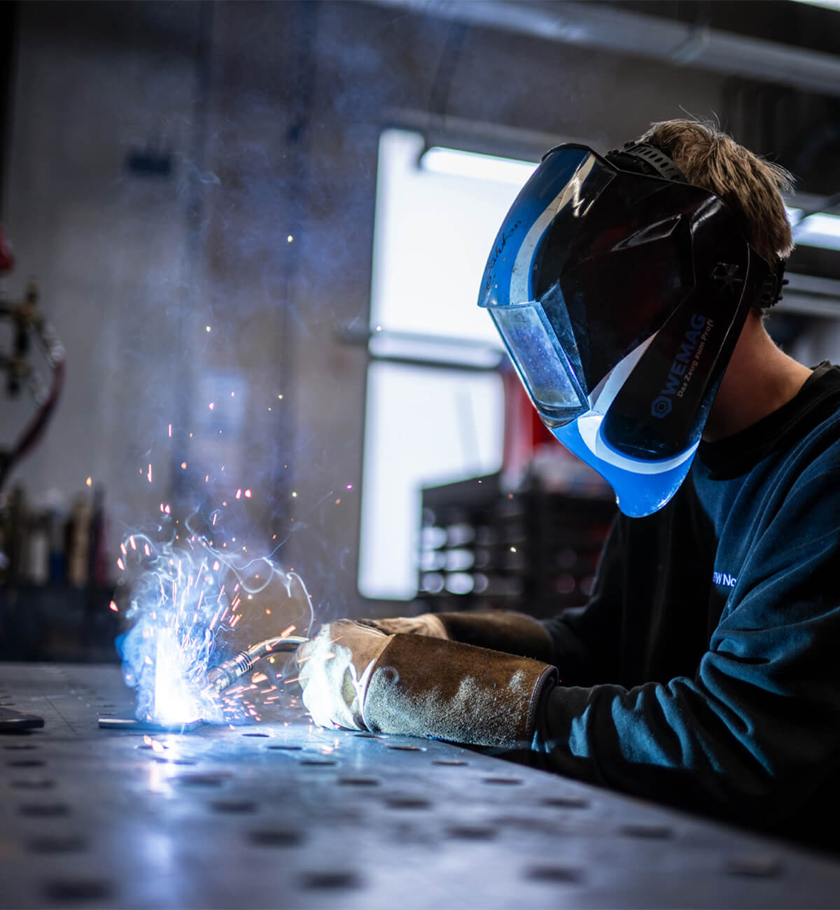 Person mit Schutzhelm und Handschuhen beim Schweißen an einer Metallplatte in einer Werkstatt