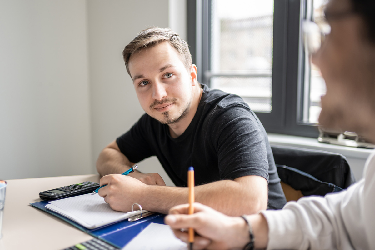 Personen sitzen an einem Tisch mit Stiften und Blöcken sowie Taschenrechnern vor sich. Eine Person schaut in die Kamera.