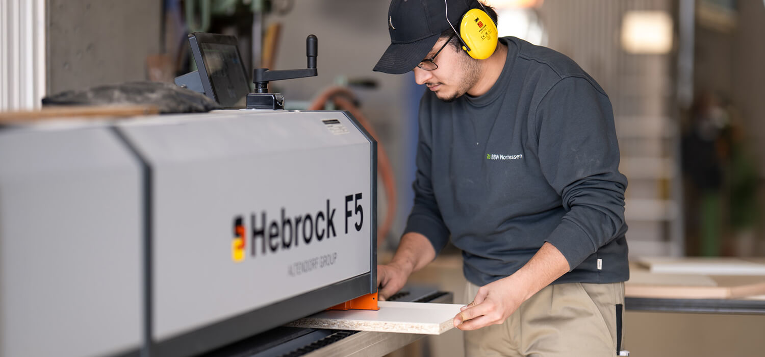 Junger Mann mit Gehörschutz bedient eine Holzbearbeitungsmaschine und führt ein Holzbrett ein