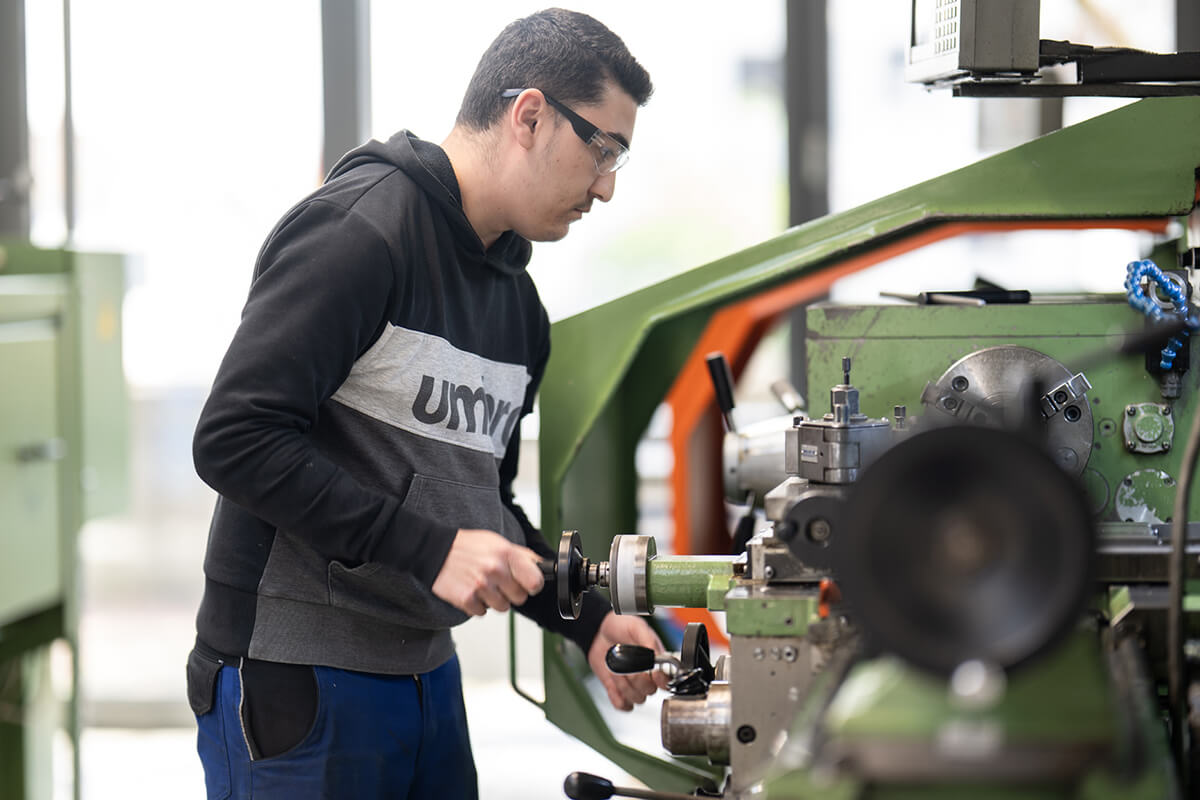 Junge Person mit Schutzbrille bedient eine grüne Metallbearbeitungsmaschine in einer Werkstatt