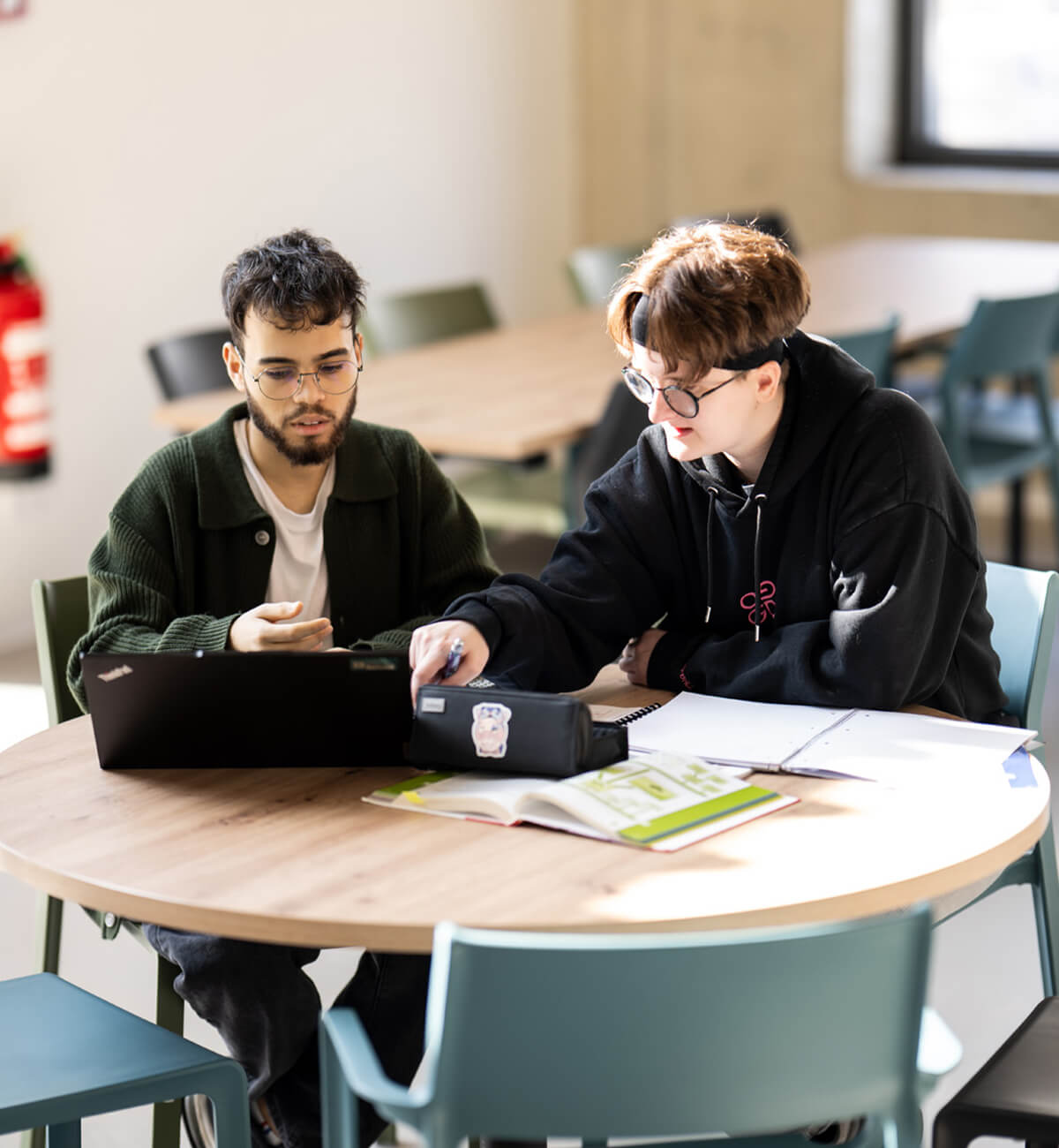 Zwei junge Menschen sitzen an einem runden Tisch und arbeiten gemeinsam mit Laptop und geöffneten Büchern