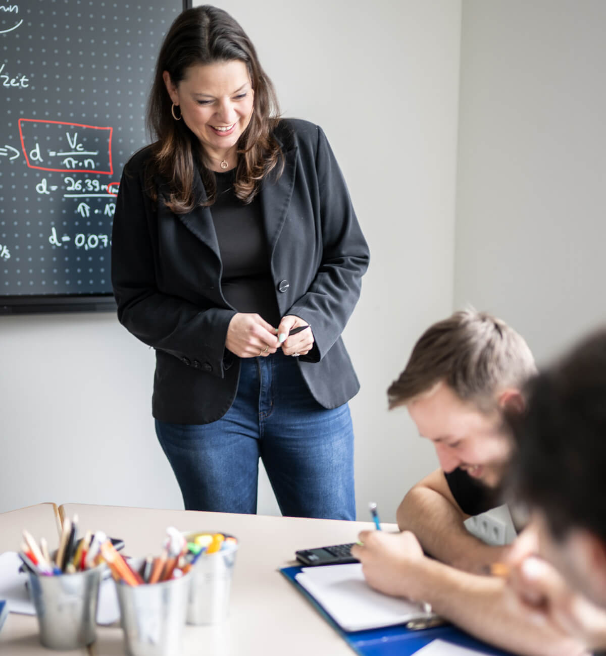 Lehrende Person steht lächelnd vor einer Tafel mit Formeln und unterrichtet zwei Schüler, die an einem Tisch schreiben