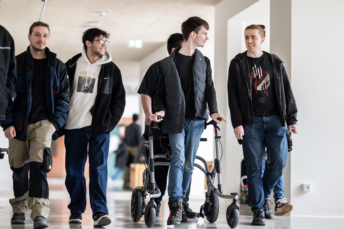 Gruppe junger Männer geht in einem hellen Flur, eine Person benutzt einen Rollator