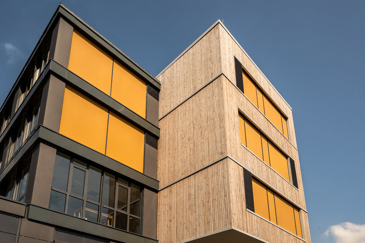 BBW-Gebäude mit Holzfassade und großen Fenstern mit gelben Sonnenschutzrollos vor blauem Himmel