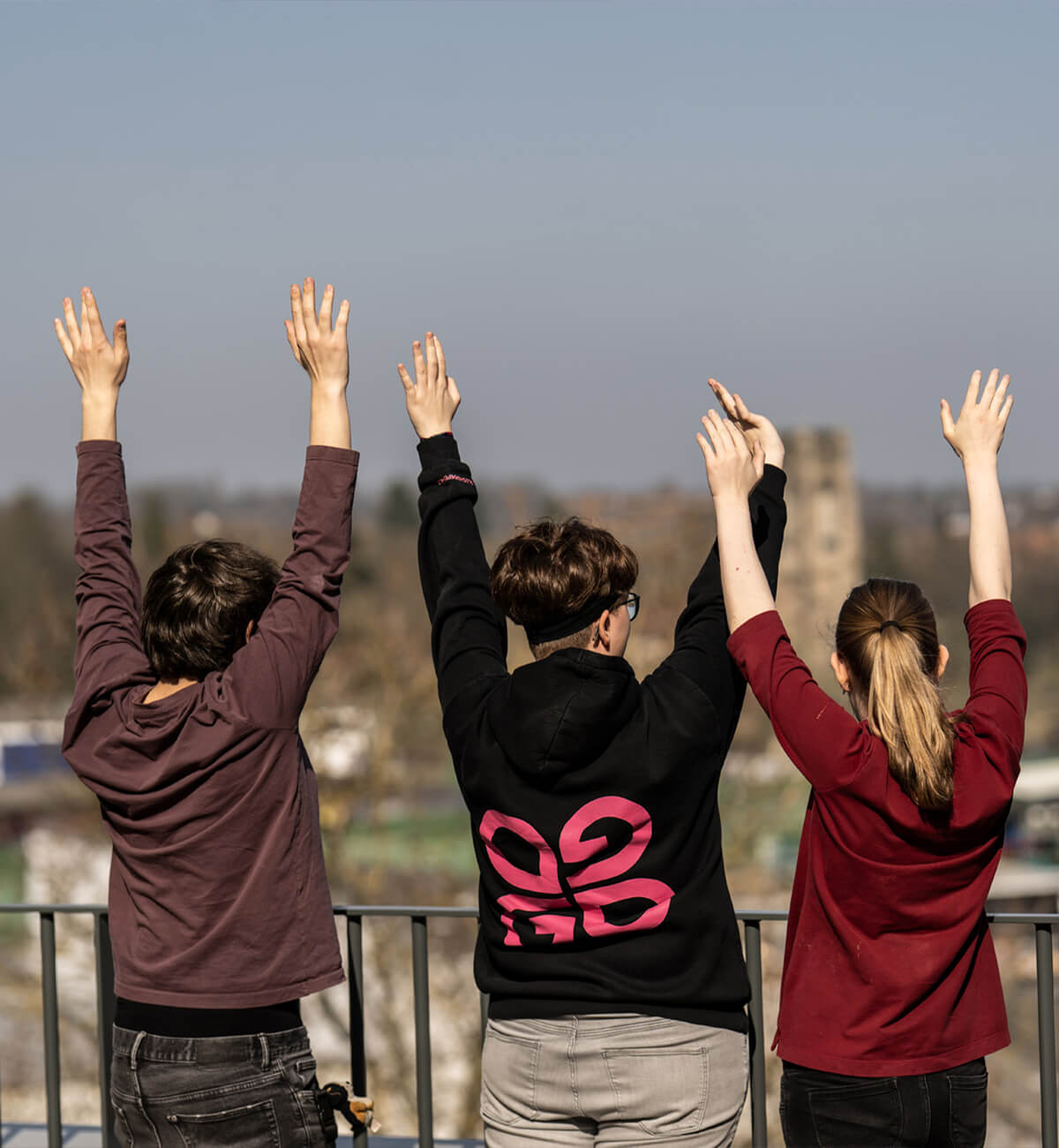 Drei Personen stehen mit dem Rücken zur Kamera und heben die Arme in die Luft, im Hintergrund unscharfe Stadtlandschaft