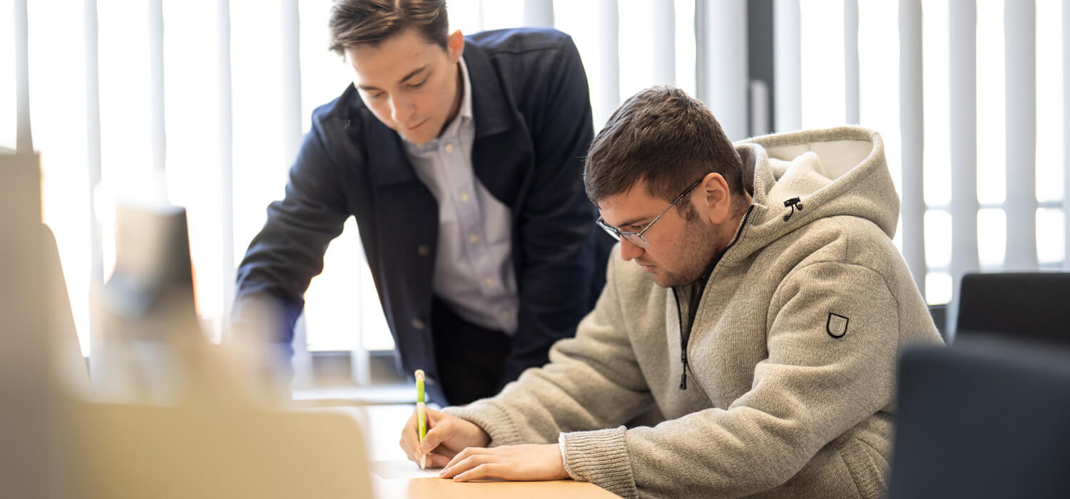 Zwei Personen n einem Büro, einer steht und zeigt auf ein Dokument, der andere sitzt und schreibt konzentriert